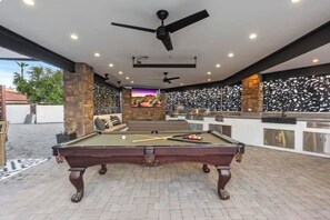 A pool table with a ceiling fan, a seating area with a view of the outdoors, and a bar area with a decorative wall. - Lux Resort - Pool, Pickleball, Hot Tub, and more! (Phoenix)