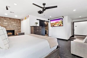 A modern bedroom with a large bed, a ceiling fan, a stone fireplace, and a TV on the wall.