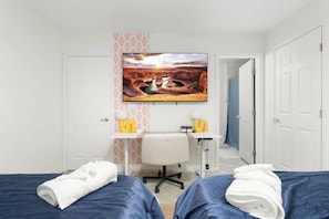 A hotel room with two beds, a desk, a chair, a painting, and a door.
