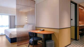 A hotel room with a bed, a desk, a chair, a small stool, and a partition.