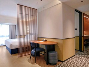 Ein Hotelzimmer mit einem Bett, einem Schreibtisch, einem Stuhl, einem kleinen Hocker und einer Raumtrennung.