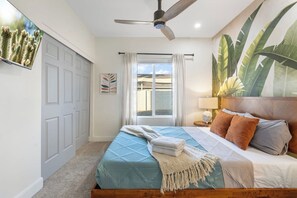 Ein Schlafzimmer mit Bett, Deckenventilator, Fenster mit Vorhängen, Wand-Fernseher und einer großen Pflanze.