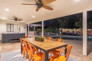 An outdoor patio with a wooden table and orange chairs, a ceiling fan, and a pool with a mural in the background. - Ultimate Backyard | Pool, Pickleball, and Golf! (Scottsdale)
