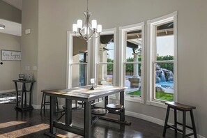 Un comedor con una mesa grande, dos taburetes de bar y una araña. Hay tres ventanas amplias que dan a un jardín con una cascada.
