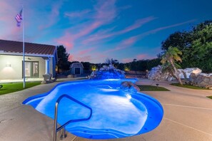 Una piscina con una cascada, rodeada de un área ajardinada con rocas y una palmera.