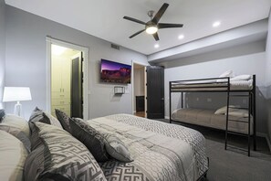 Ein modernes Hotelzimmer mit Etagenbett, einem großen Bett mit gemusterten Bettwäsche, einem Deckenventilator und einem Bild an der Wand.