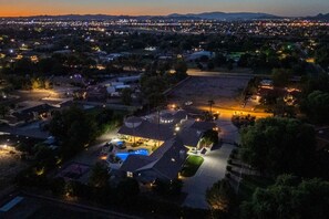 An aerial view of a residential area with houses, a swimming pool, and a well-lit driveway at dusk. - Pickleball Main House | Pool | Hot Tub | Arcade (Peoria)