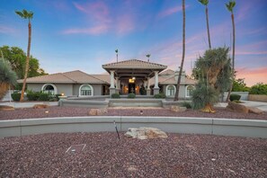 A two-story house with a tiled roof, a covered front porch, and a landscaped front yard with rocks and palm trees. - Pickleball Main House | Pool | Hot Tub | Arcade (Peoria)