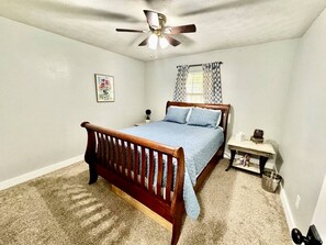 Ein Schlafzimmer mit einem hölzernen Bett, einem Deckenventilator, einem kleinen Nachttisch und einem Fenster mit Vorhängen.