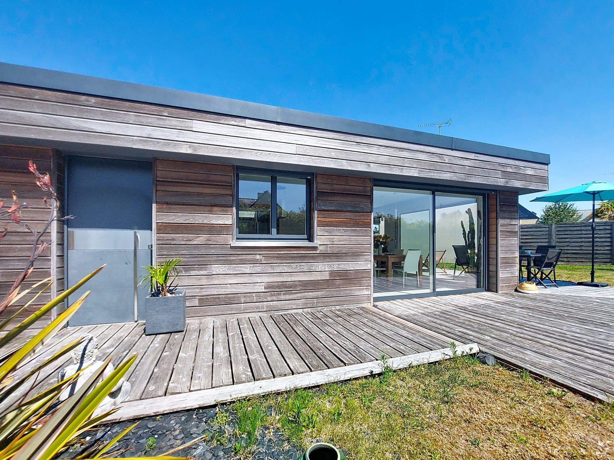 Ein modernes Haus mit einer Holzterrasse, großen Schiebeglasfenstern und einer kleinen Sitzgelegenheit im Freien.