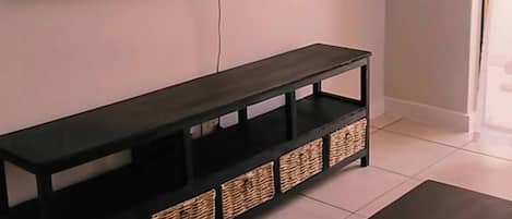 A living room with a flat-screen TV mounted on the wall, a dark wooden bench with storage baskets, and a coffee table.