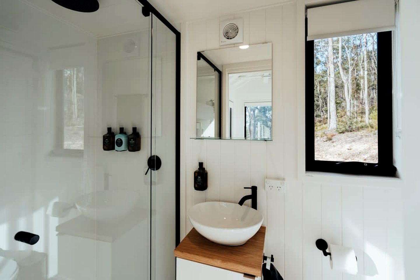 A bathroom with a glass shower door, a white sink with a wooden countertop, a mirror above the sink, and a window with a forest view.