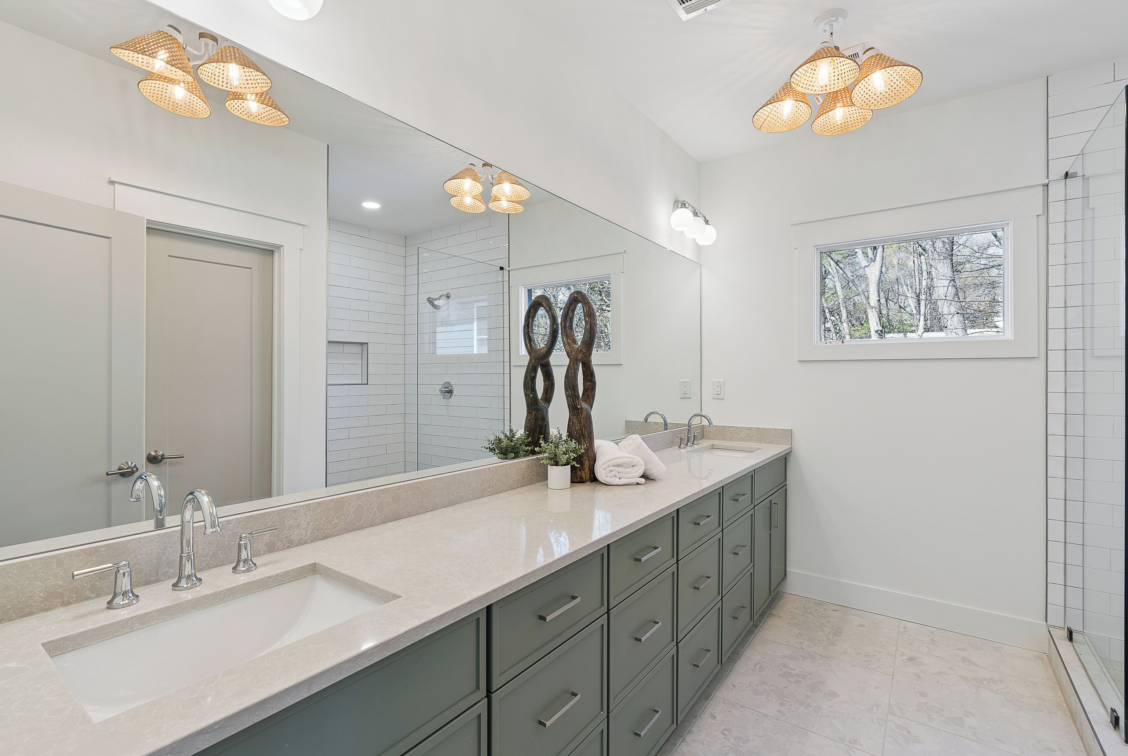 A modern bathroom with a large mirror, two sinks, green cabinets, and a window with a view.