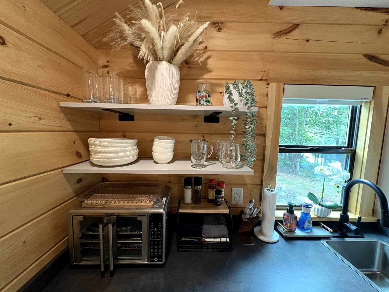 A compact kitchen with wooden walls, a built-in oven, and a window with a view of greenery.