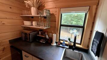 A compact kitchen with wooden walls, a window with a view, and basic amenities like a sink, microwave, and storage shelves.