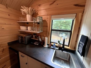 Une cuisine compacte aux murs en bois, dotée d’une fenêtre offrant une belle vue et équipée des commodités essentielles, comme un évier, un four à micro-ondes et des étagères de rangement.