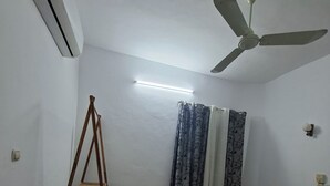 Una habitación con cama, ventilador de pie, ventilador de techo, estante y ventana con cortinas.