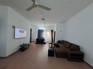 Ein Wohnzimmer mit Deckenventilator, einem an der Wand befestigten Fernseher, einem braunen Sofa und einem Essbereich mit Tisch und Stühlen.