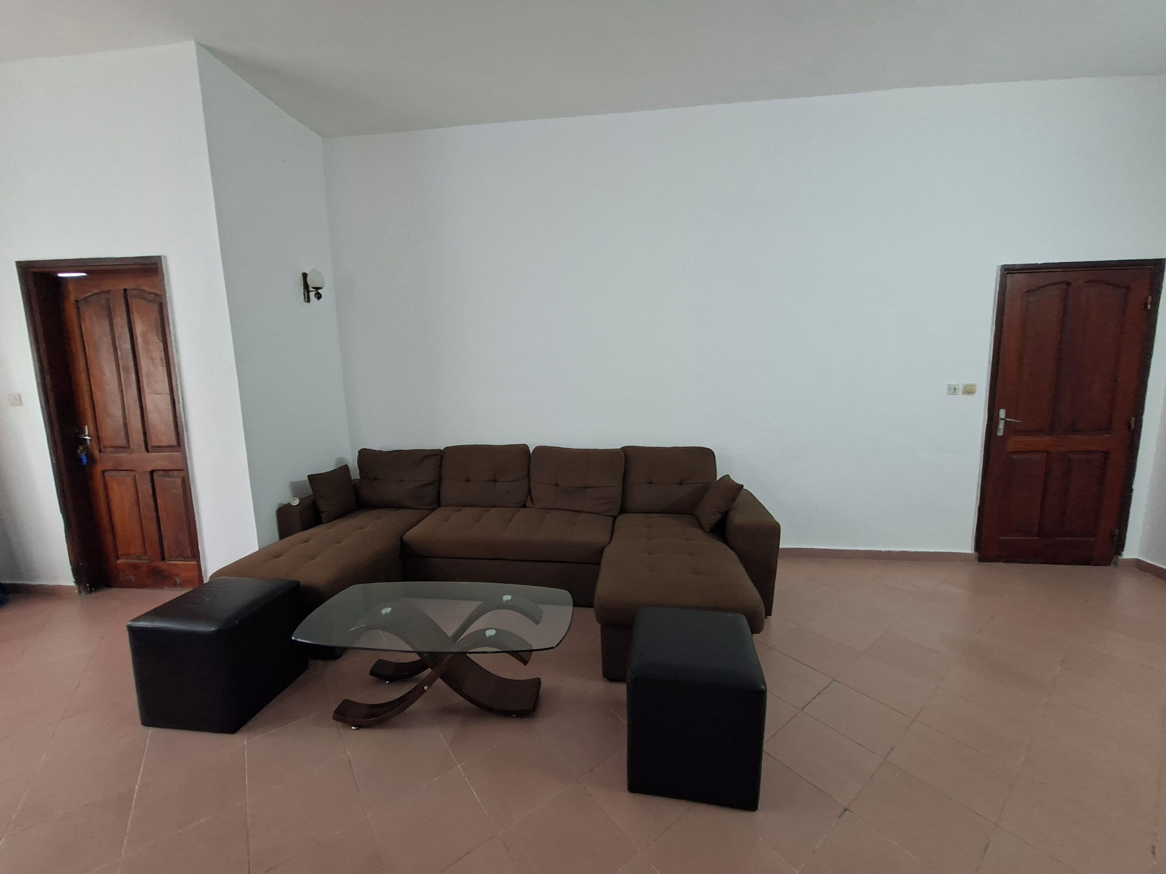 Un salon comprenant un canapé d’angle marron, une table basse avec un plateau en verre et deux poufs noirs.