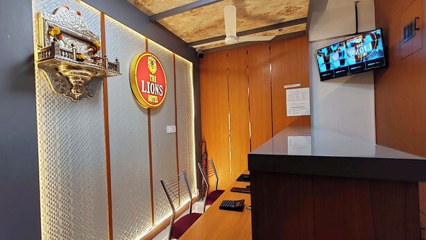 Ein Hotel-Empfangsbereich mit einer Rezeption, einem an der Wand montierten Fernseher, einer Wand mit einem Löwenwappen und einem gemusterten Fenster.