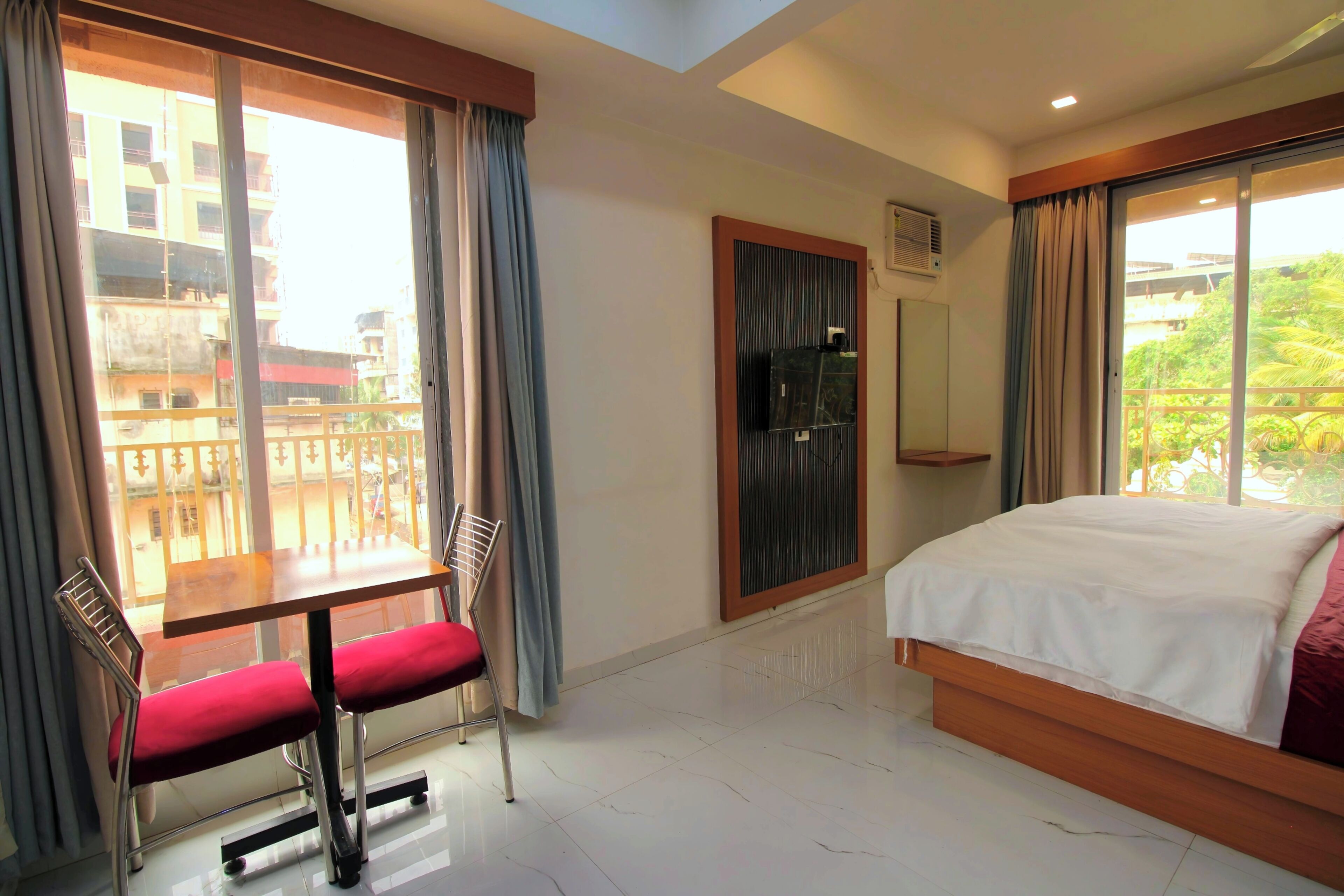 A hotel room with a bed, a table with a chair, a window with curtains, and a view of buildings outside.
