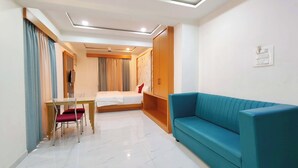 Habitación de hotel con cama, sofá azul, mesa de comedor con sillas y televisor montado en la pared.