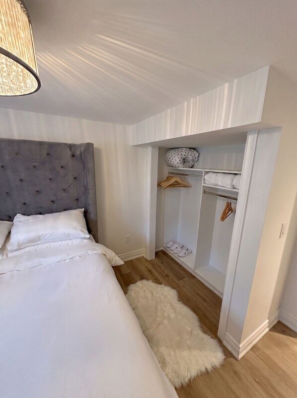 Une chambre avec un lit, une tête de lit grise à motifs, un tapis blanc et moelleux, une armoire avec espace pour suspendre les vêtements et un porte-chaussures.