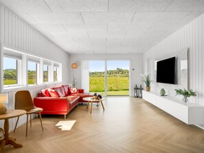 A spacious living room with a red sofa, wooden flooring, large windows, and a flat-screen TV mounted on the wall. - "Tameran" -  from the sea by Interhome (Hals)