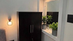 Ein Zimmer mit einem dunklen Kleiderschrank, einem kleinen Nachttisch, einem Wandregal mit Topfpflanzen und einer Wandleuchte.