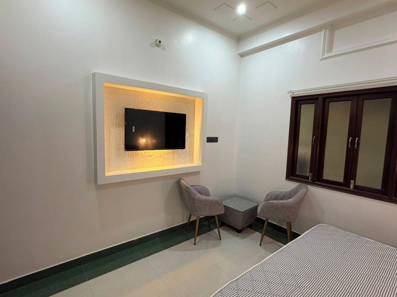 Una habitación con una televisión de pantalla plana montada en la pared, dos sillas, una mesita y una cama.