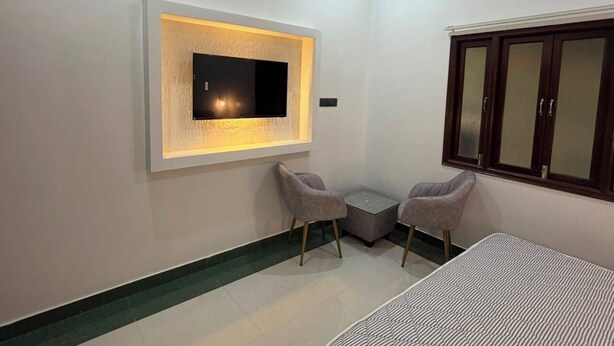 Ein Zimmer mit Flachbildfernseher an der Wand, zwei Stühle, einem kleinen Tisch und einem Bett.