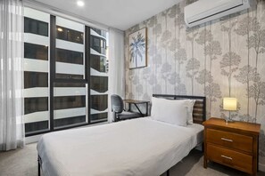 A hotel room with a bed, a desk, a chair, a nightstand, and a large window with a view of buildings. - High-Rise 2-Bed Apt at Trio Boxhill with Car Park (Box Hill)