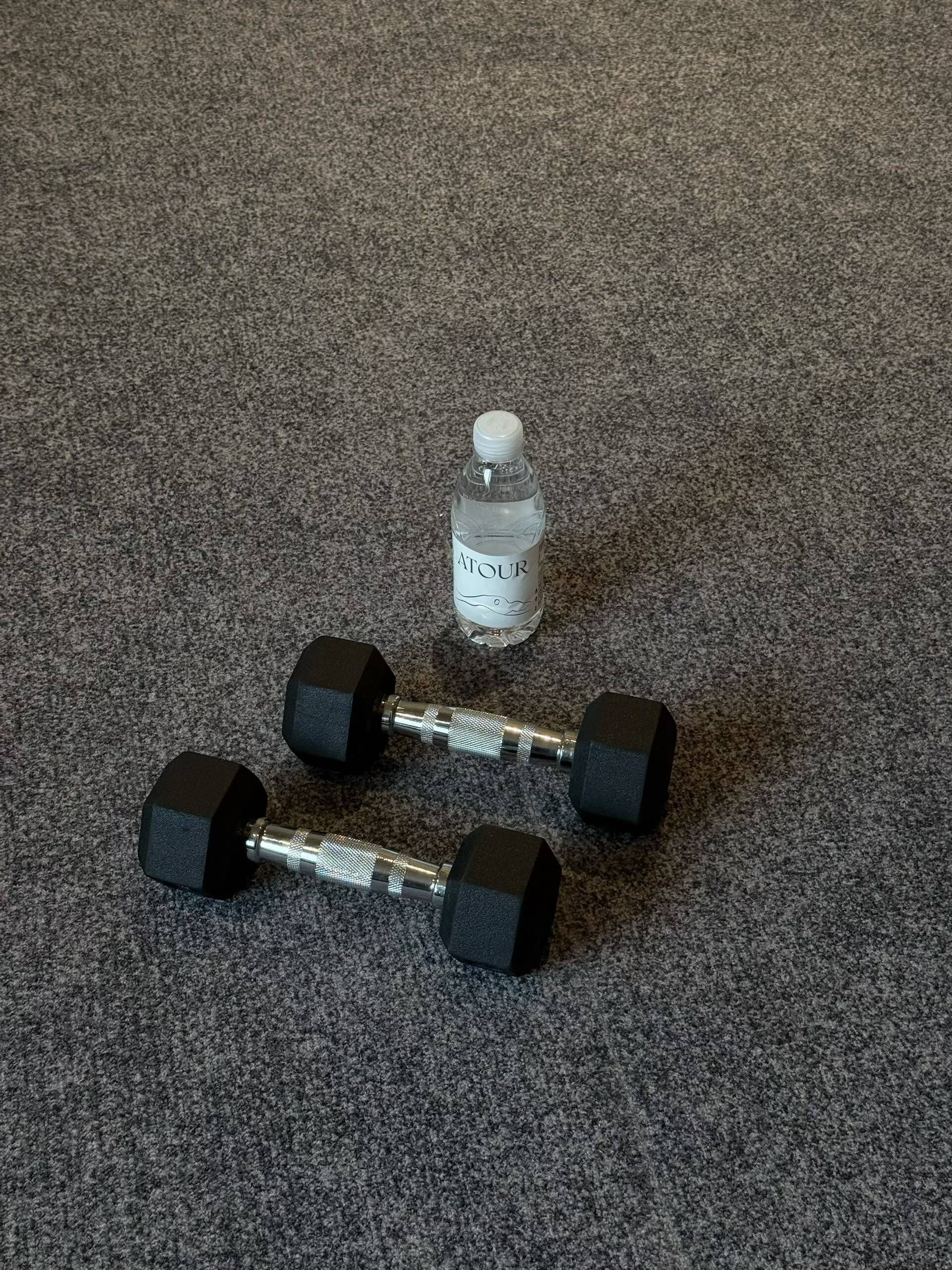A pair of black dumbbells and a clear water bottle with the label 'ATOUR' on a textured carpet.