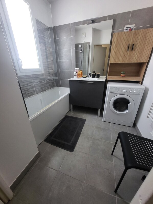 A compact bathroom with a bathtub, a sink, a mirror, a washing machine, and a small shelf with toiletries. - Luminous Cocoon - All-Around Comfort - Sleeps 4 to 6 (Montpellier)