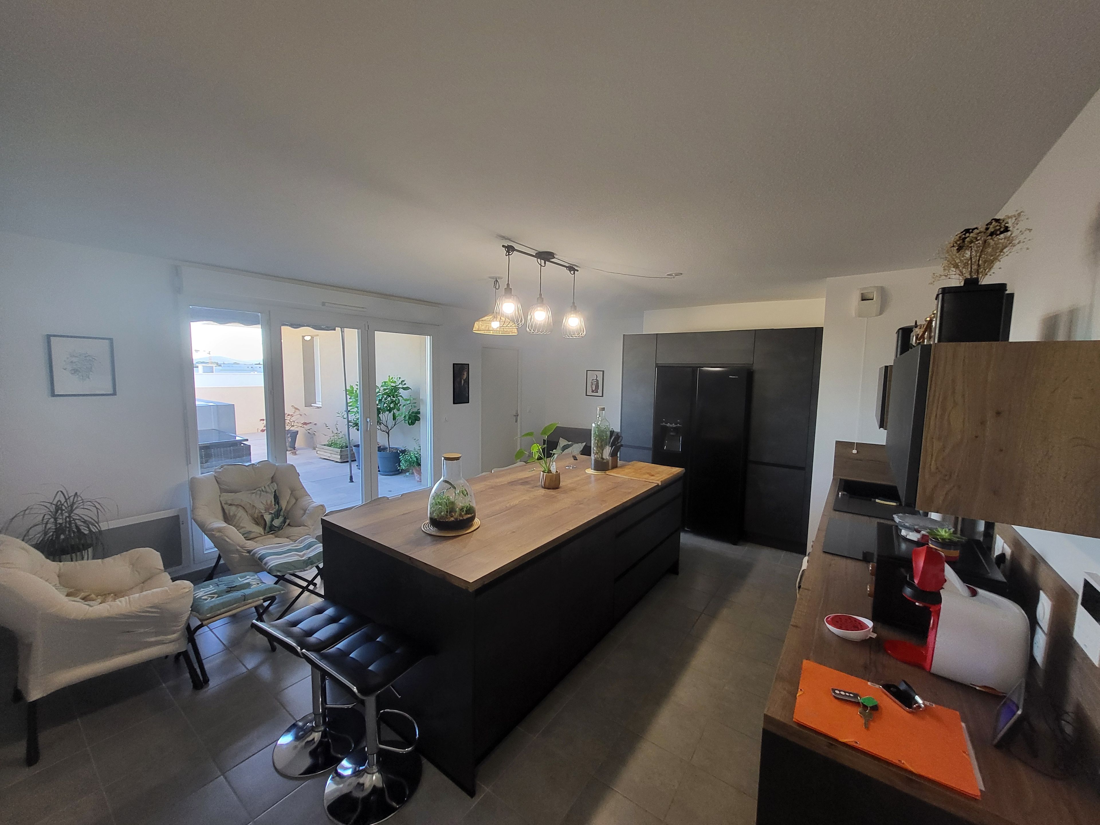 A modern kitchen with a central island, a dining area with a sofa and bar stools, and a view of the outdoors through a sliding door.