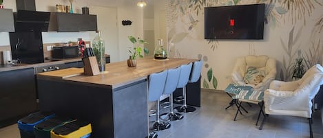 A modern kitchen with a central island, bar stools, a flat-screen TV, and a dining area with a chair.