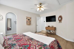 Un dormitorio con cama, ventilador de techo, televisor, espejo y puerta.