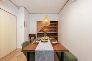 Un comedor con una mesa de madera, sillas verdes, una lámpara colgante y una estantería al fondo.