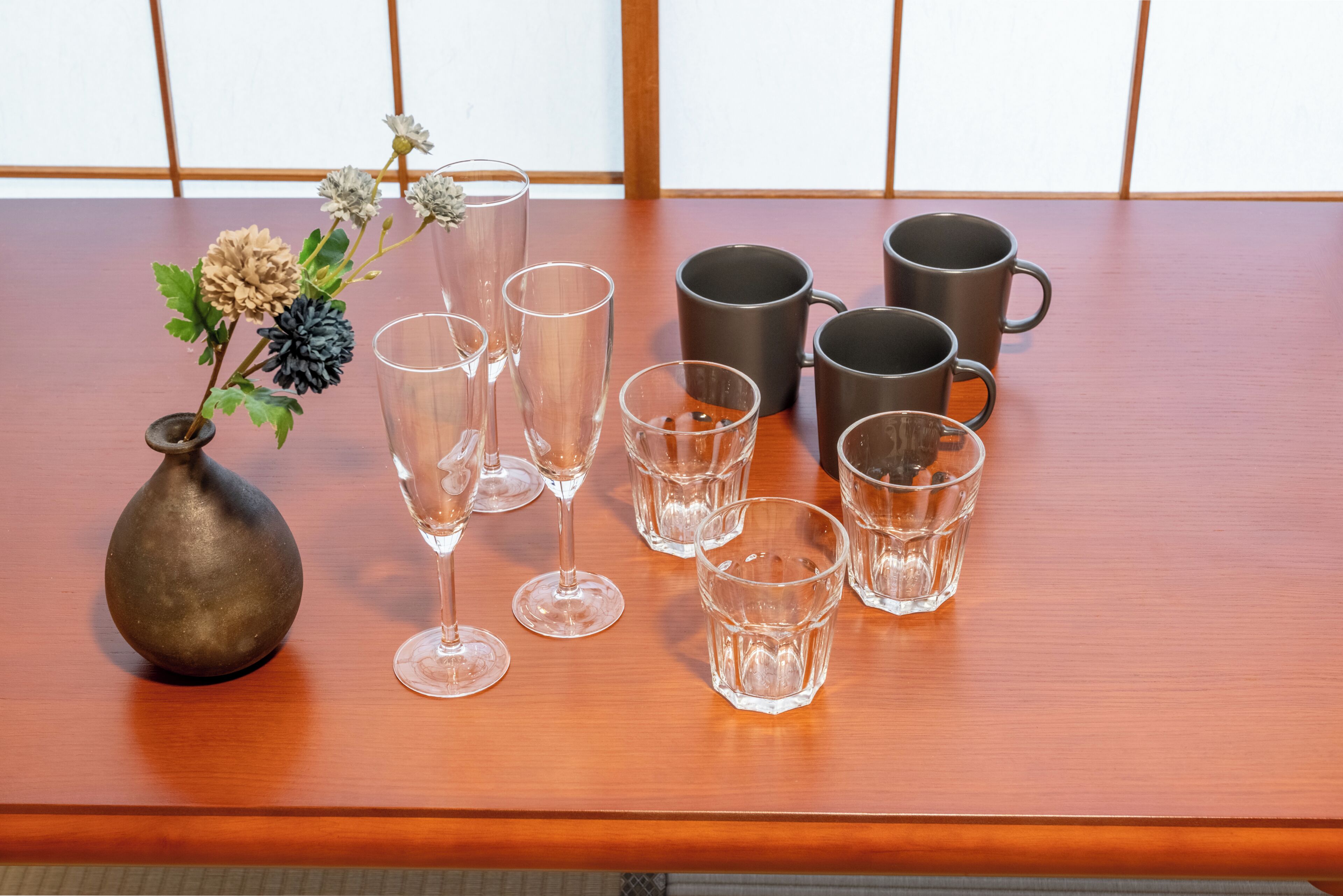Una taula de fusta amb un ram de flors, diversos vasos de cristall i un conjunt de tasses negres.