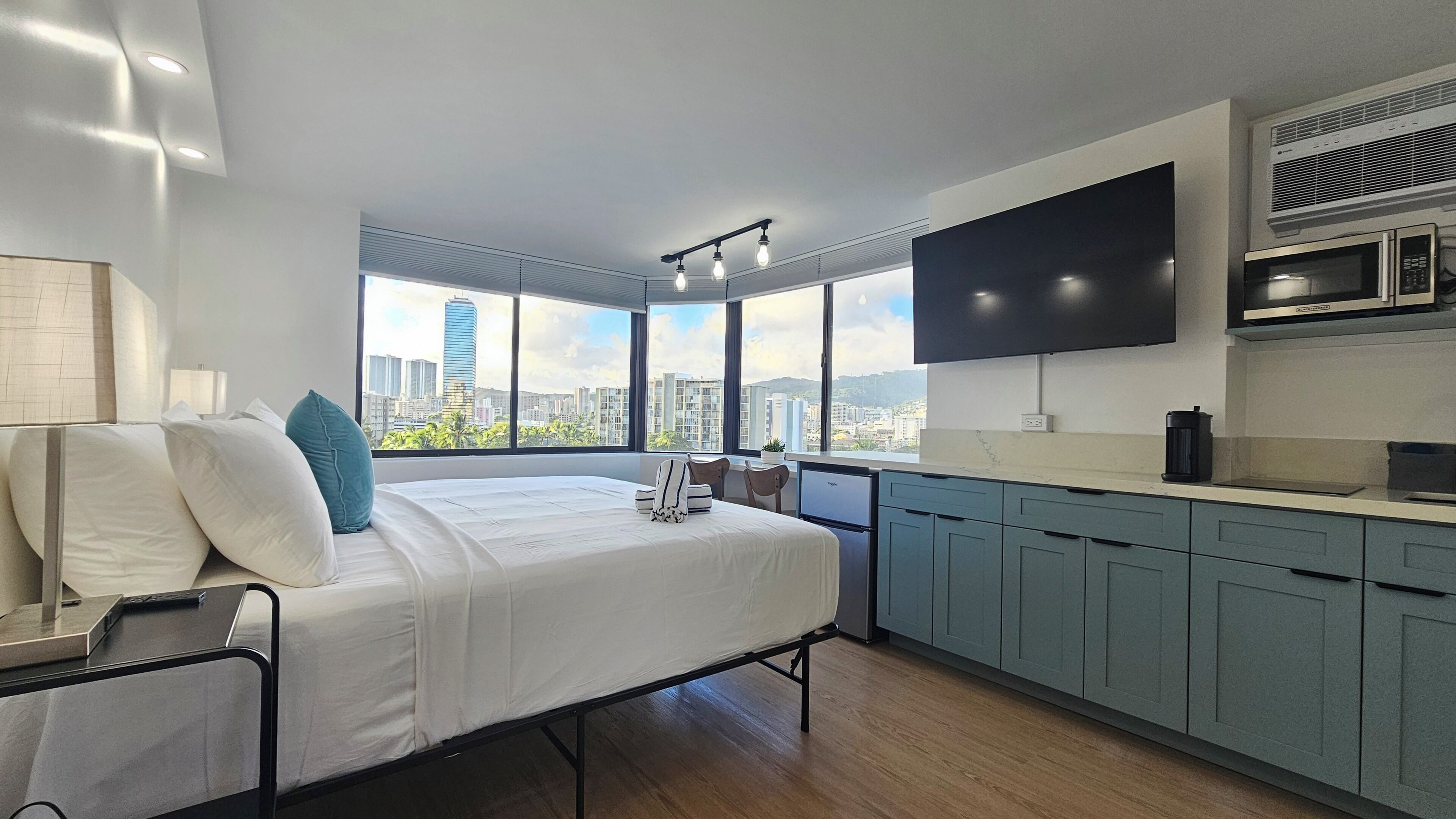 A modern hotel room with a large bed, a city view, a flat-screen TV, and a kitchenette.