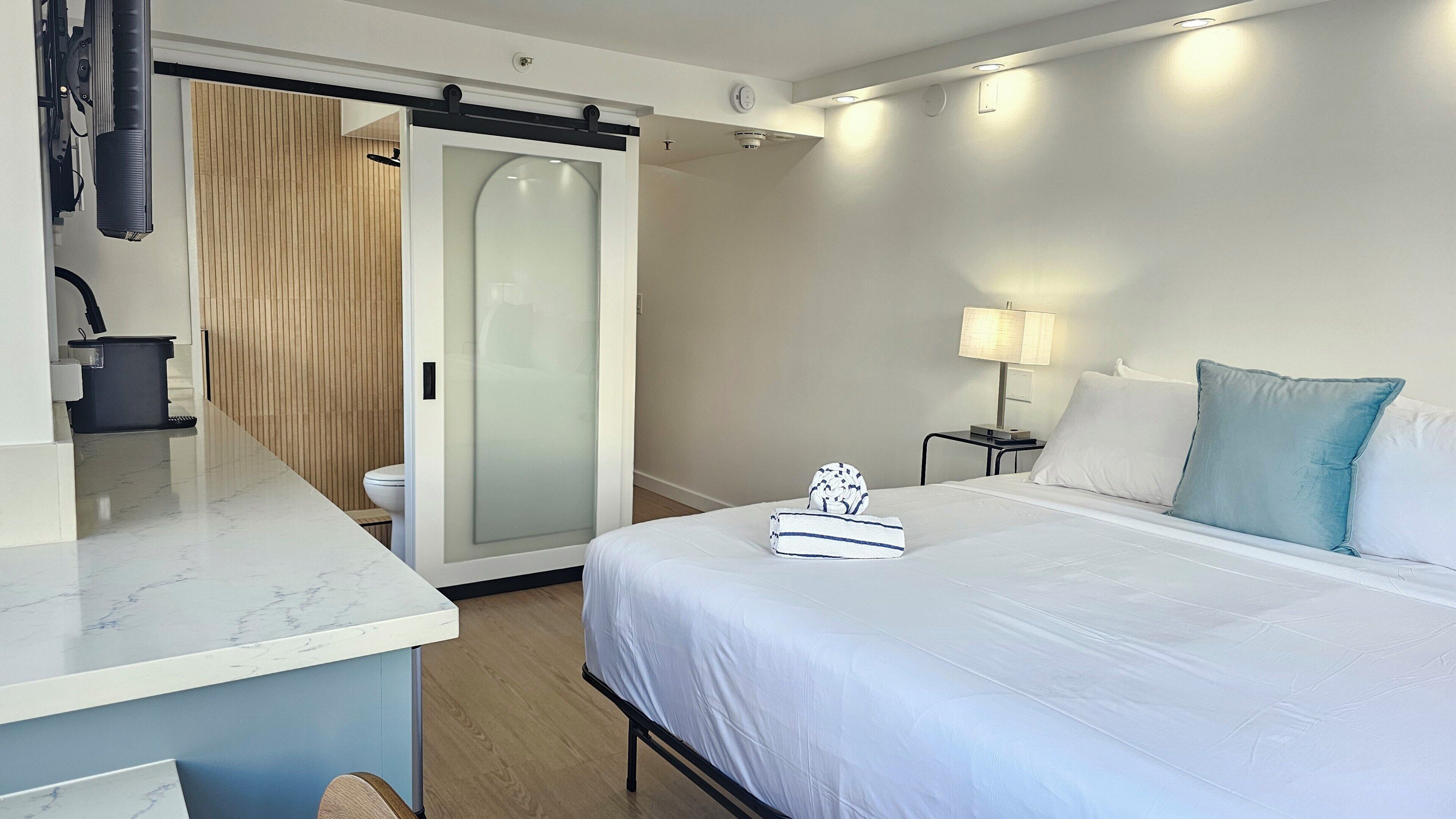Habitació d'hotel amb un llit, una taula de nit, una làmpada, una tovallola i una porta corredissa que dona a un bany.