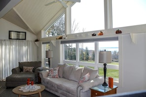A living room with a grey sofa, a smaller grey armchair, a round wooden coffee table, and two floor lamps. - Charming Waterview Cottage with Sunset Views & Private Deck Near Acadia National Park | Ellsworth, Maine (Ellsworth)