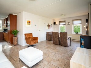 Ein modernes Wohnzimmer mit einer Couch, Sesseln und einem weißen Hocker. Es gibt eine Küchenzeile mit Holzschränken und einem Topf mit Pflanze.