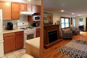 A kitchen with wooden cabinets, a stove, and a microwave. There is a fireplace with a mantel and two patterned armchairs.