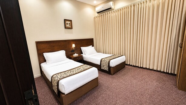 A hotel room with two beds, a nightstand with a lamp, a picture on the wall, and a window with curtains. - Dome Hotel Service Apartments (Riyadh)