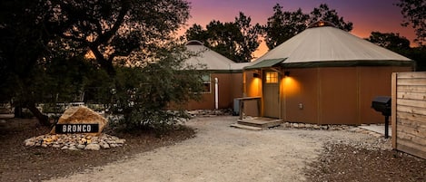 A two-story yurt with a gravel driveway, a wooden sign labeled 'BRONCO,' and a wooden fence.