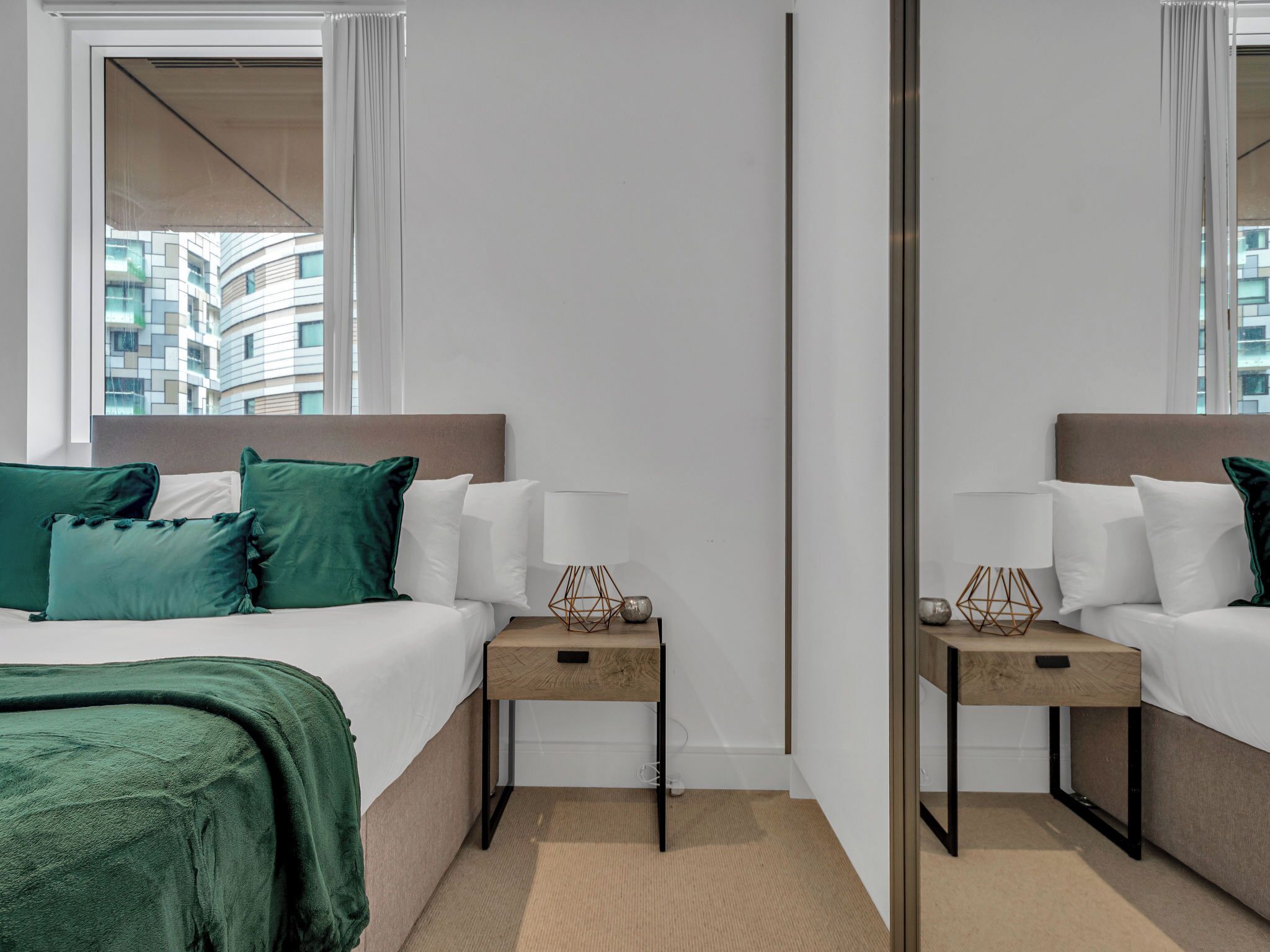 A hotel room with a bed, bedside tables, a mirror, and a view of buildings.