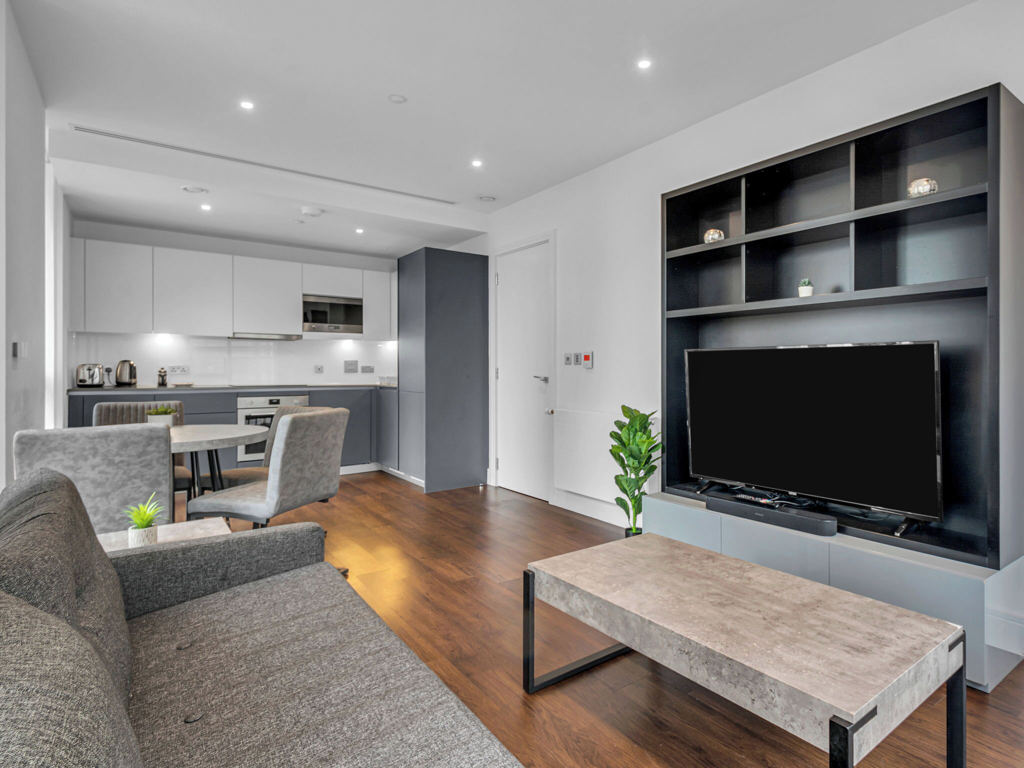 A modern living room with a flat-screen TV, grey sofas, a coffee table, and a kitchen in the background.