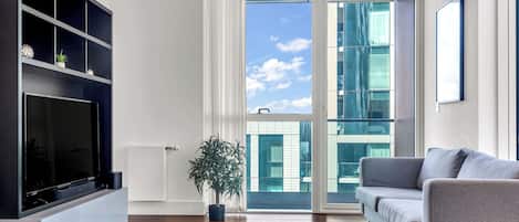 A modern living room with a flat-screen TV, a grey sofa, a potted plant, and a large window with a view of buildings.
