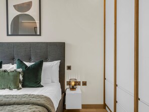 A modern bedroom with a grey upholstered headboard, white bedding, and a bedside table with a lamp. - Stratford Loft by Interhome (London East)